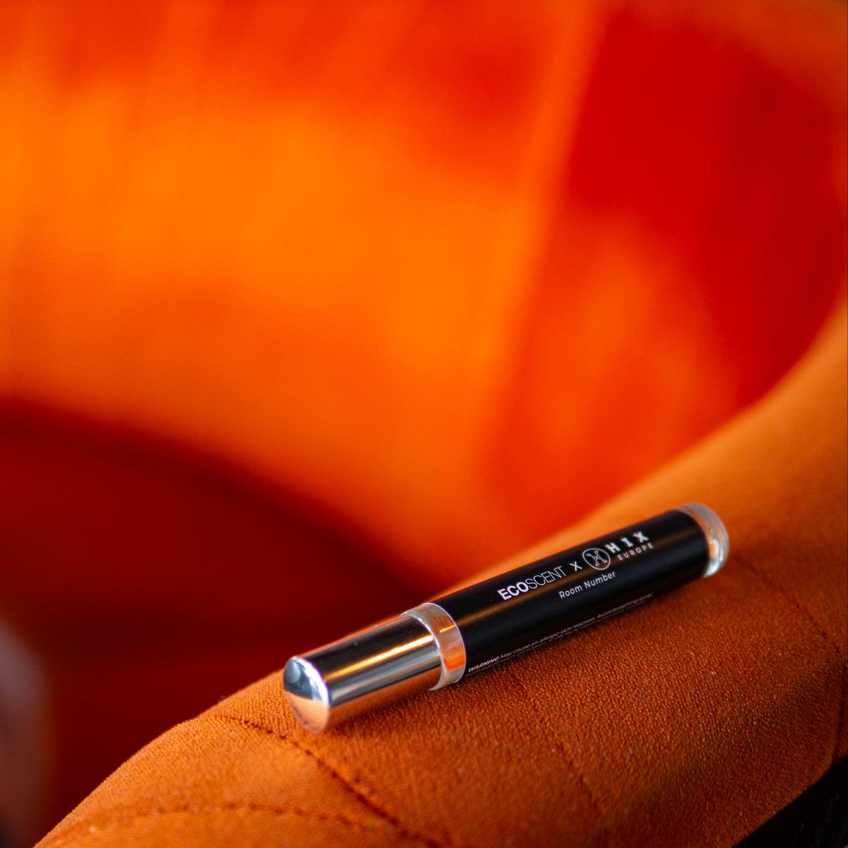 A black and silver cosmetic EcoScent room spray resting on the arm of a textured orange chair, with a warm orange background.