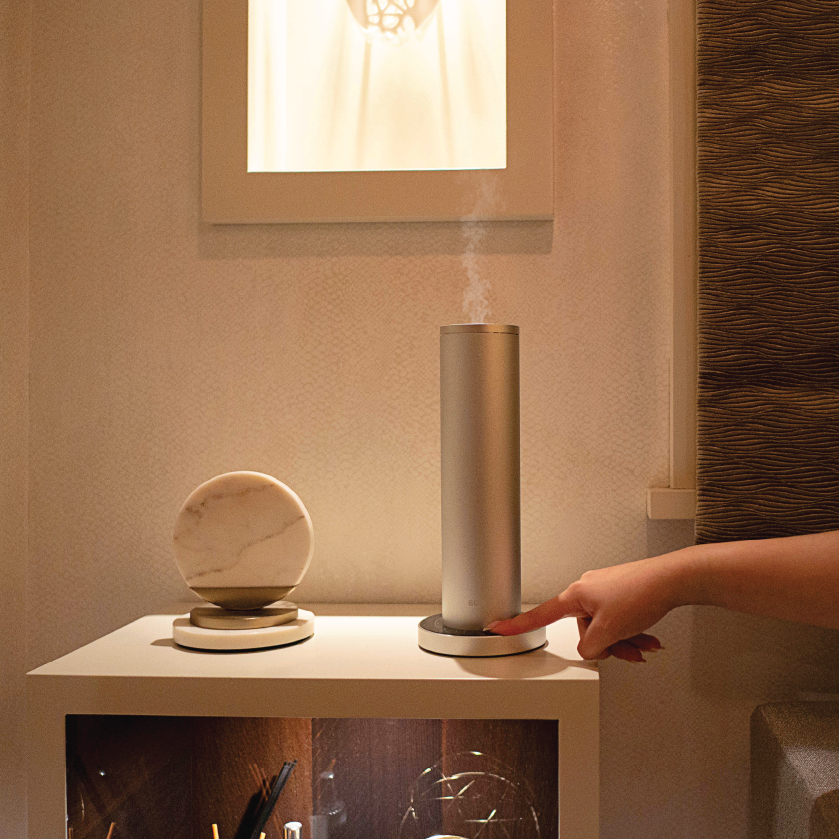 Hand using a small cylindrical scent machine emitting a fine mist on a table with decorative items.