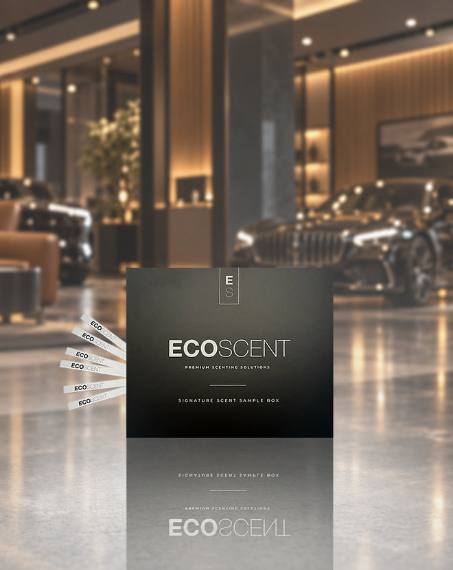 A dark charcoal ECOSCENT premium defining fragrance sample box stands in a luxury car showroom. Several white sample cards are fanned out to the left, and a blurred car and modern interior are visible in the background.