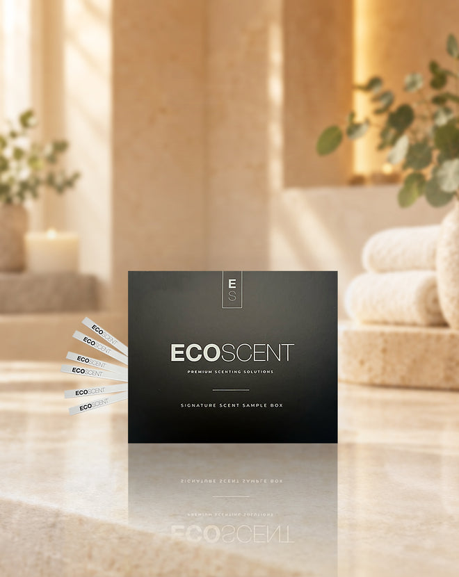 A black EcoScent premium scenting solution box stands on a polished stone surface in a warm, softly lit, spa-like setting with folded towels and greenery in the background. Several white sample strips are fanned out to the left.
