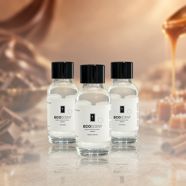 Three small, clear glass bottles of EcoScent fragrance oil have black caps and white labels. They are arranged on a reflective surface. A warm, amber background sits behind them. Honey drips in the distance.