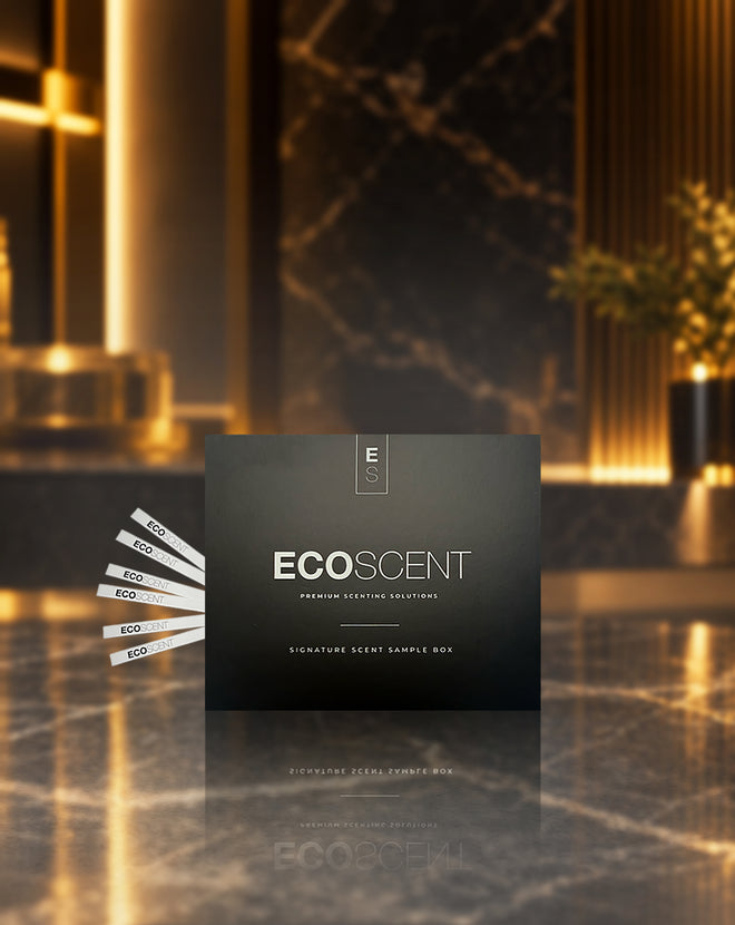 A dark charcoal grey square product box labelled EcoScent sits on a polished marble surface with a blurred luxury interior in the background. Several white sample strips are fanned out to the left of the box.