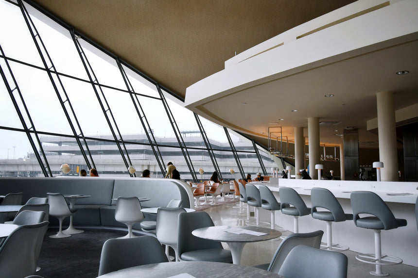 A modern airport lounge with curved architecture, large slanted windows, gray chairs, round tables, and people seated in the background. The view outside shows part of the airport runway and terminal structures.