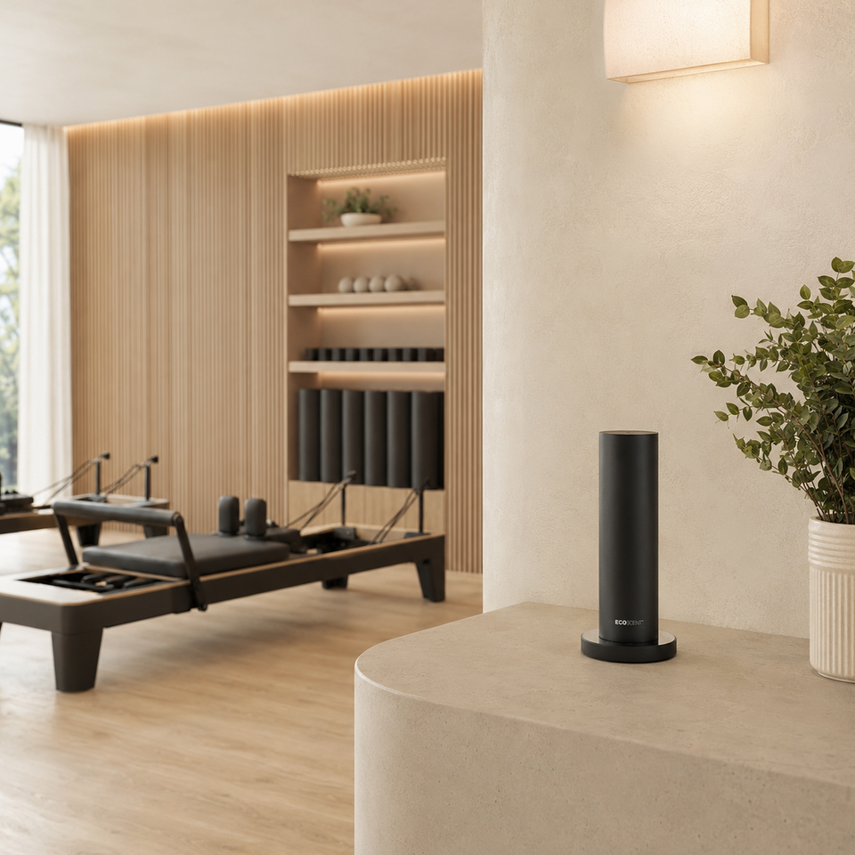 A sleek black cylindrical scent machine sits on a light beige stone counter in a modern, minimalist wellness studio with wooden reformer machines and warm ambient lighting.
