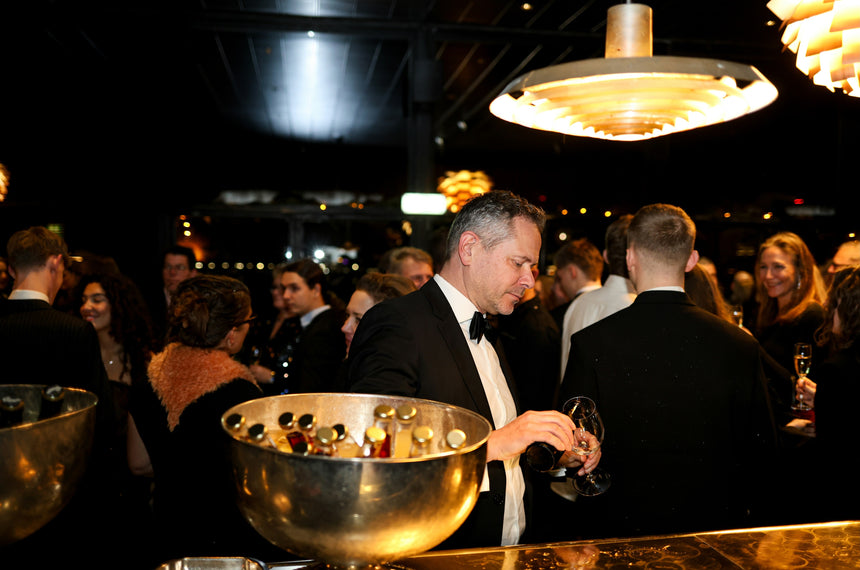 A man in a tuxedo pours a drink at a bar during a crowded, elegant evening event. People in formal attire are socializing in the background under warm, modern lighting.