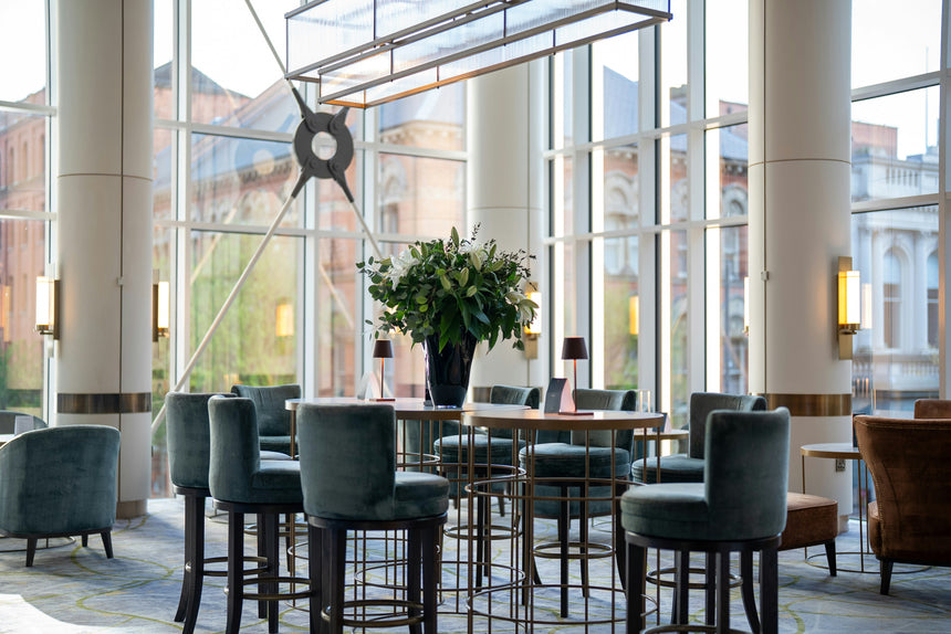 Modern, airy lounge with tall windows, teal velvet chairs around high tables, a large plant centerpiece, and city buildings visible outside. Warm lighting creates a cozy atmosphere.