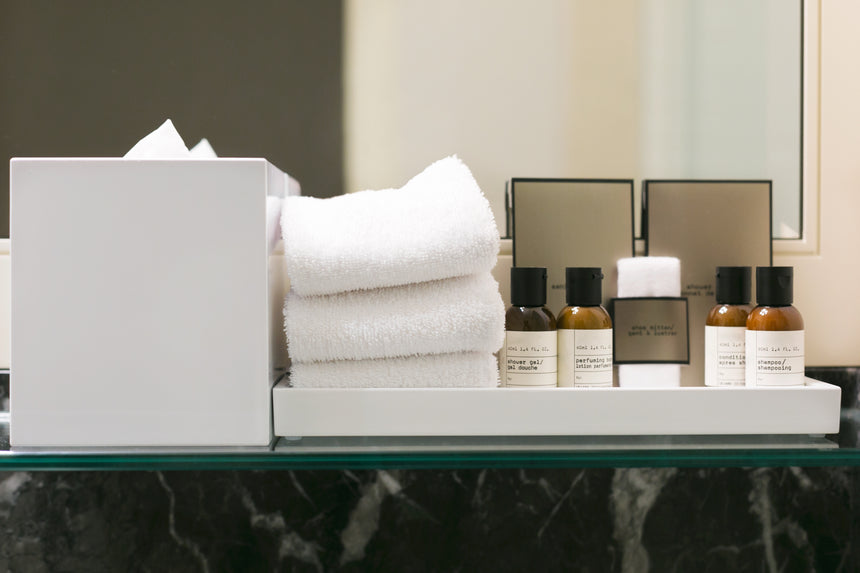 A tray with neatly folded white towels, small bottles of toiletries, tissue box, and vanity kits sits on a glass shelf above a dark marble countertop in a bathroom.