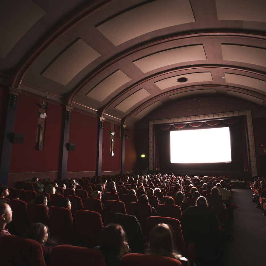 A large movie theater auditorium filled with seated audience members facing a bright white screen, with red seats, dark walls, and an arched ceiling.