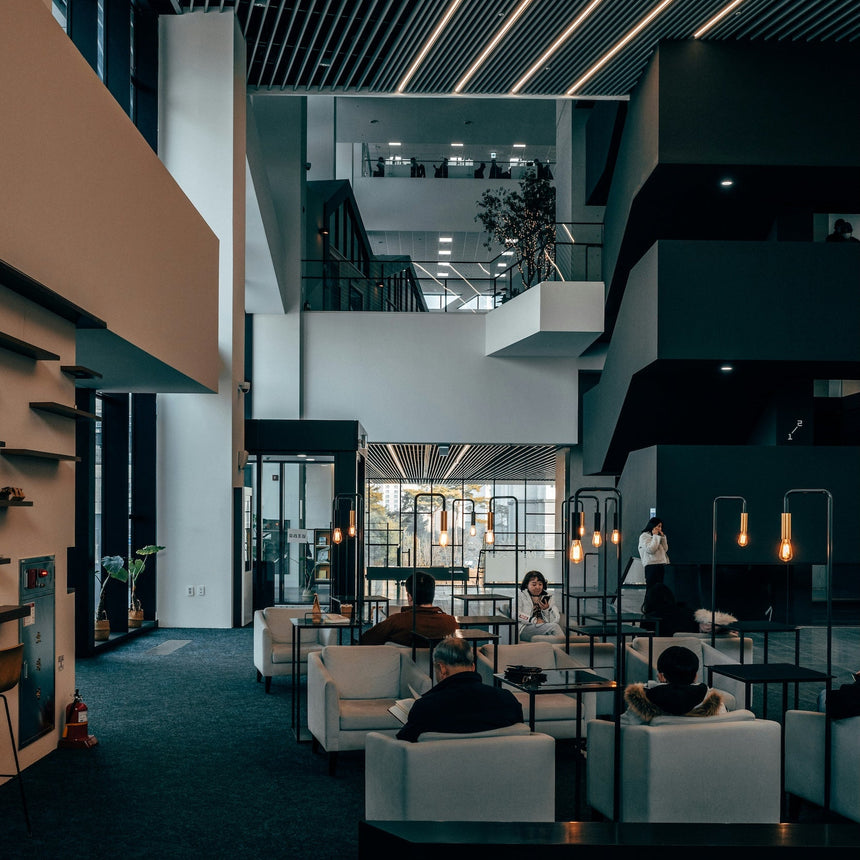 A modern multi-level office lobby with dark wood slatted ceiling, linear LED lighting, white and black seating, and several people sitting and standing in the space.
