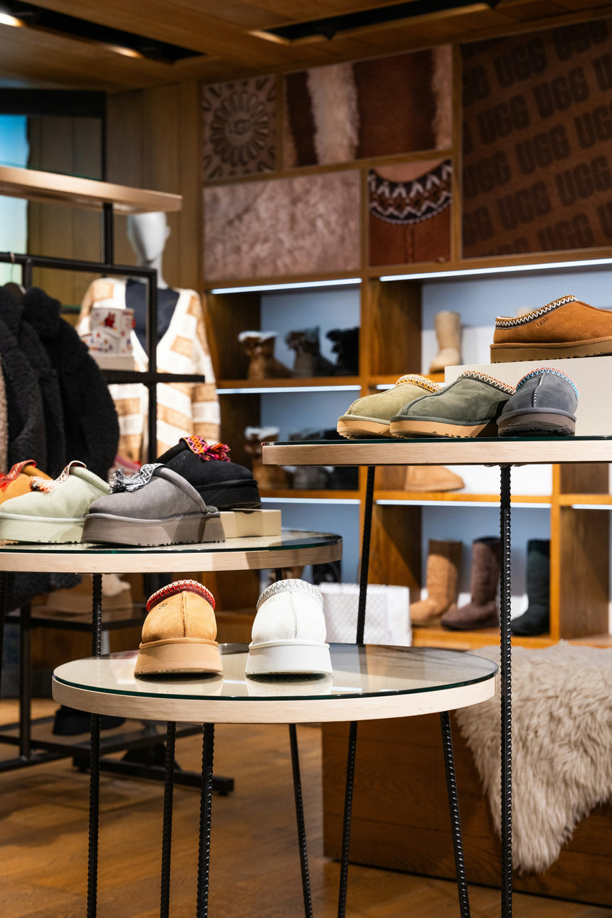 Display of UGG footwear on three circular tables, featuring pairs of slippers in various colours including grey, beige, and white against a backdrop of wooden shelves and textured wall panels.
