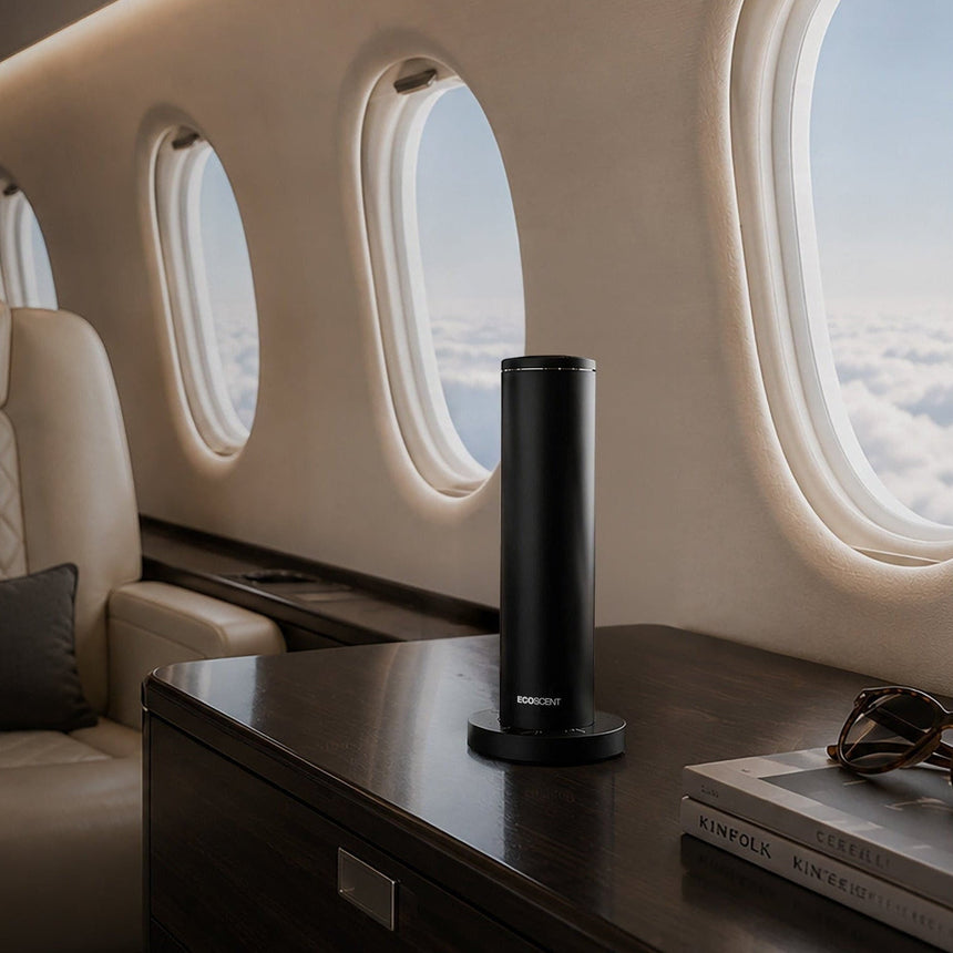 A sleek black cylindrical portable scent machine by EcoScent sits on a dark wooden console table inside a luxury private jet cabin, with cream leather seats, oval windows, and a book and sunglasses nearby.