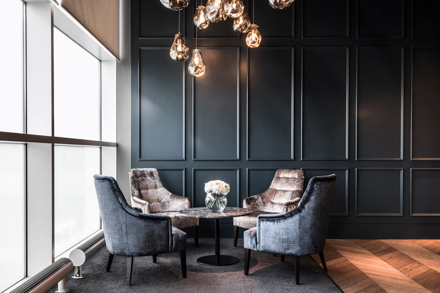 Two gray velvet chairs sit around a small round table with a vase of white flowers, under modern pendant lights in a stylish room with dark paneled walls and large windows.