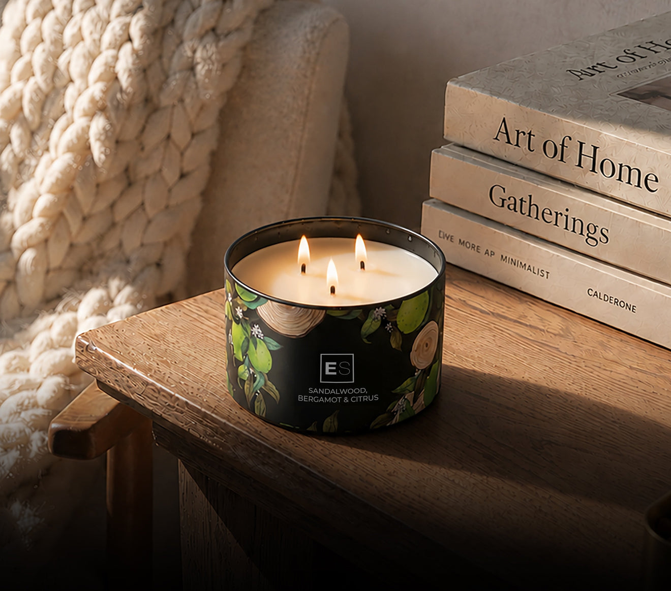 A lit black glass candle tin with a green botanical label sits on a wooden table beside stacked books and a chunky cream knit blanket.