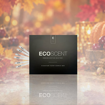 A black ECOSCENT signature scent sample box is centered against a warm, blurred autumn background with fall leaves, pumpkins, and candles. Several white sample strips labeled ECOSCENT are fanned out to the left.