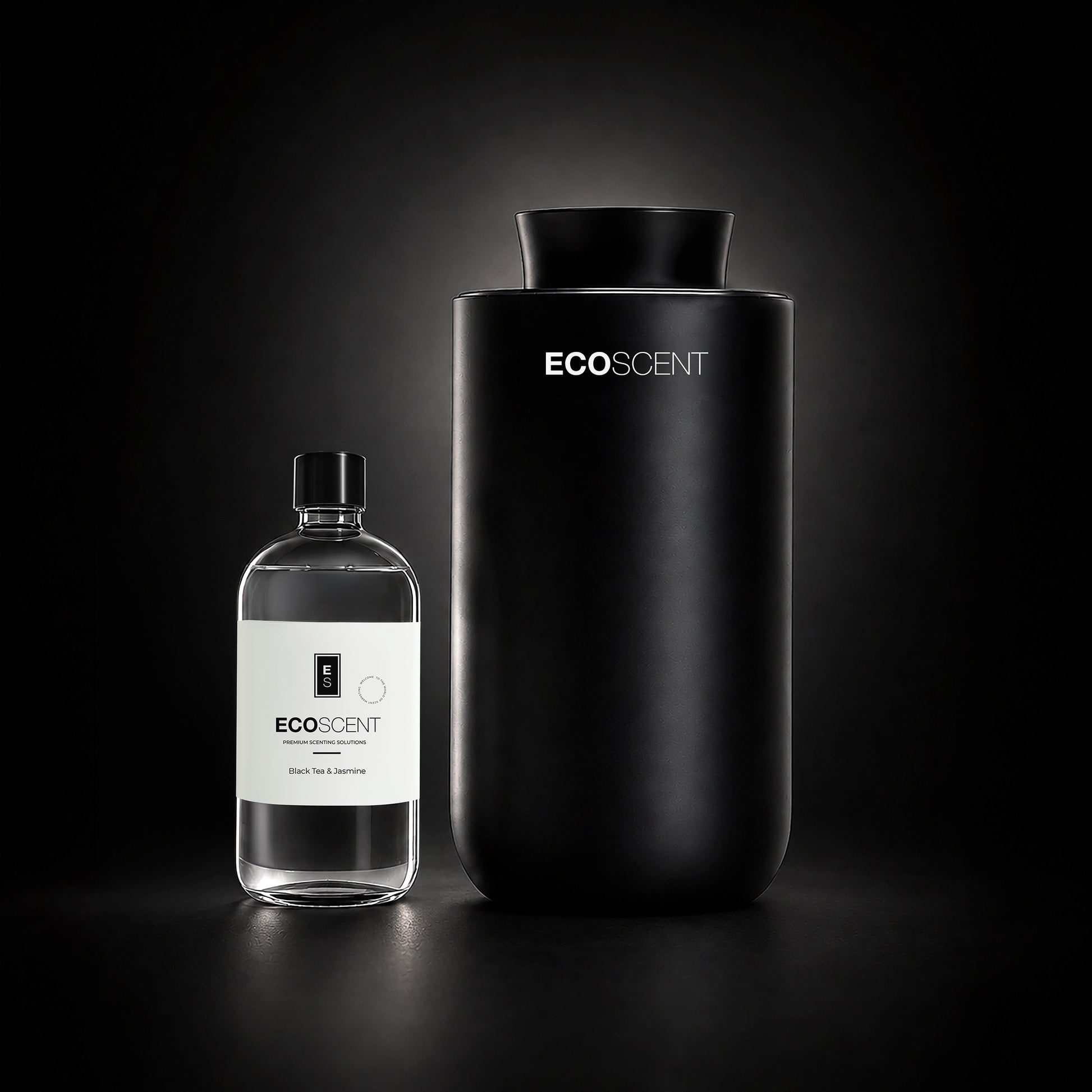 A matte black cylindrical EcoScent diffuser stands next to a clear glass bottle with a white label on a dark studio background.