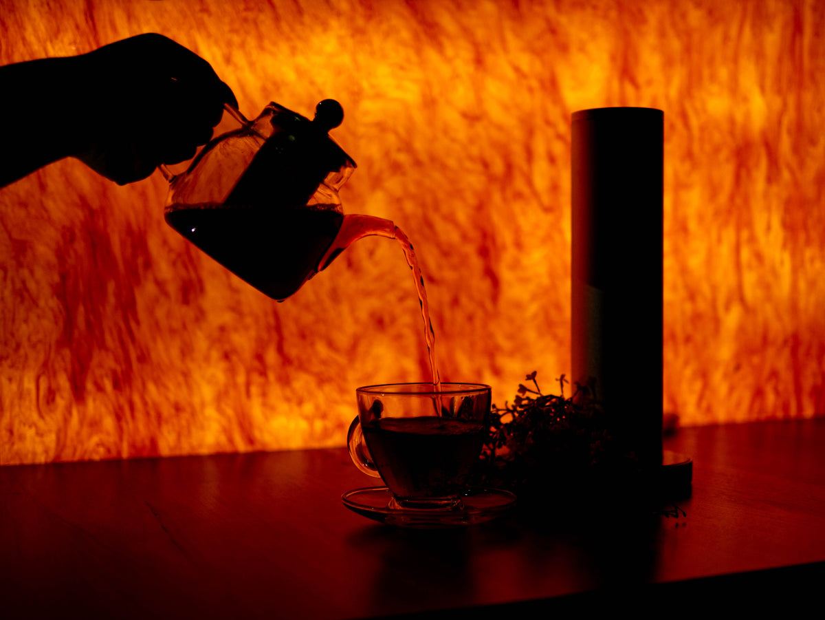 A hand silhouetted against a glowing orange textured background pours hot water from a glass teapot into a glass cup on a saucer, with a dark cylindrical container and small plant sprigs on the table.
