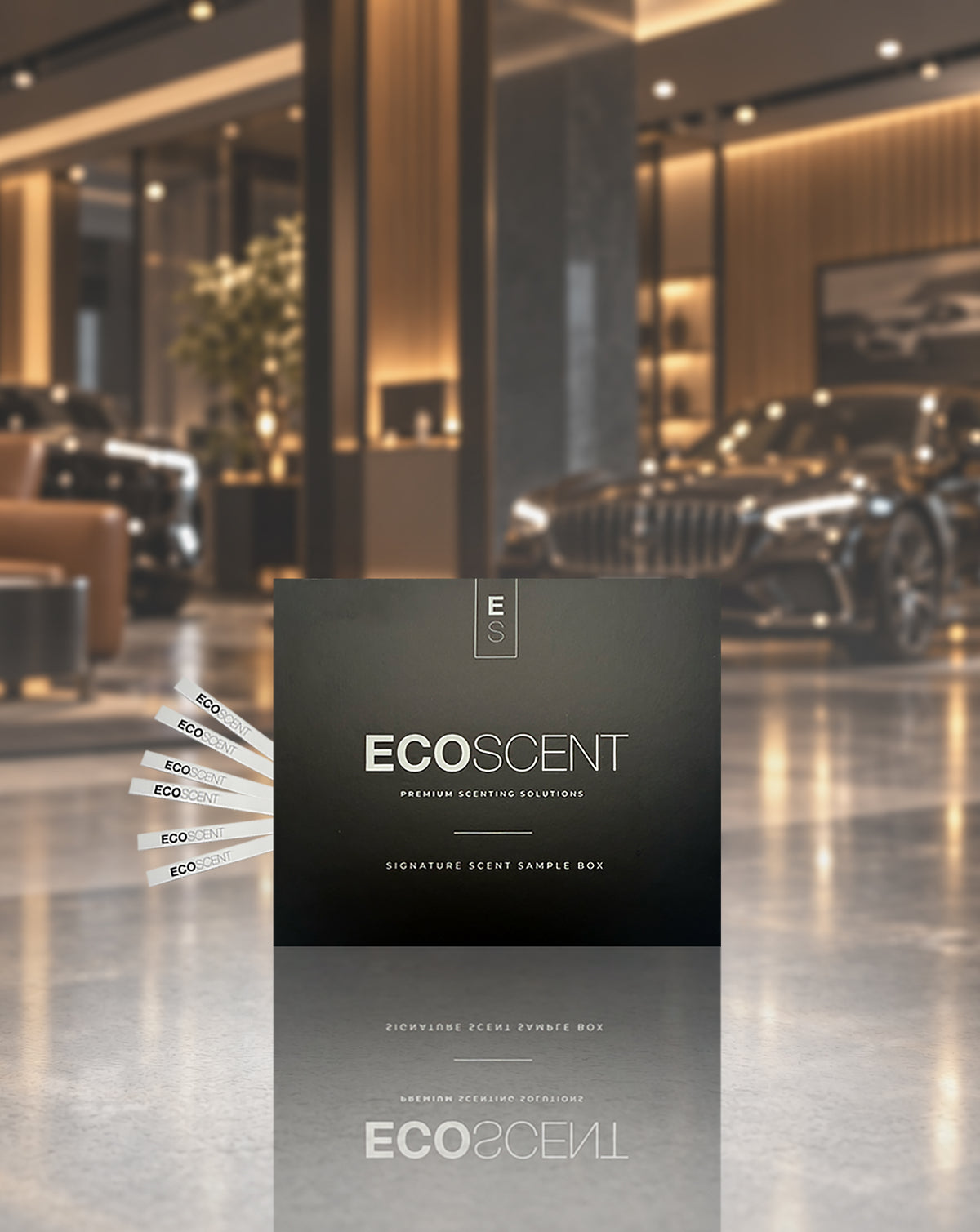 A dark charcoal ECOSCENT premium defining fragrance sample box stands in a luxury car showroom. Several white sample cards are fanned out to the left, and a blurred car and modern interior are visible in the background.