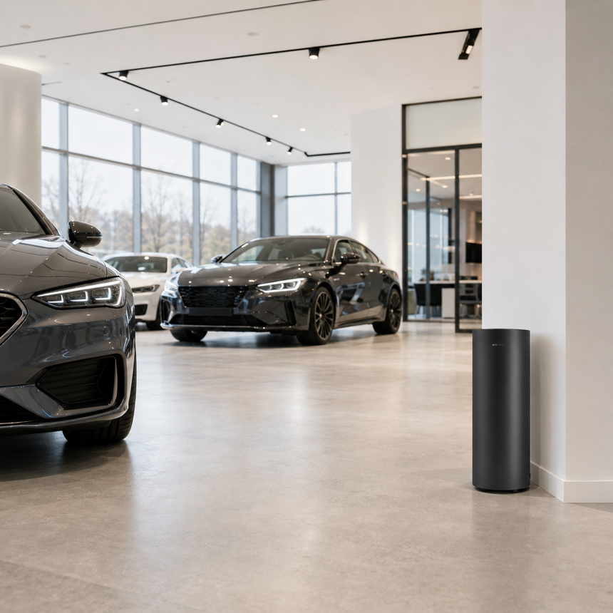 A sleek black sports car and a luxury sedan displayed in a modern car showroom with large windows and bright lighting. A cylindrical black object stands on the floor in the foreground.