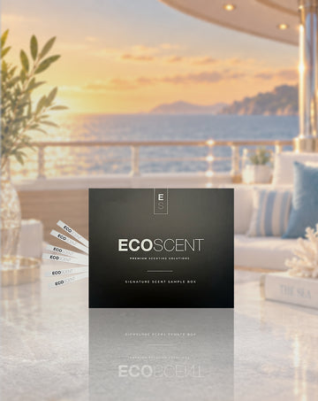 A black ECOSCENT premium setting solution sample box stands on a reflective marble surface in a luxurious outdoor patio setting at sunset, with a sea view in the background and several sample cards fanned out to the left.