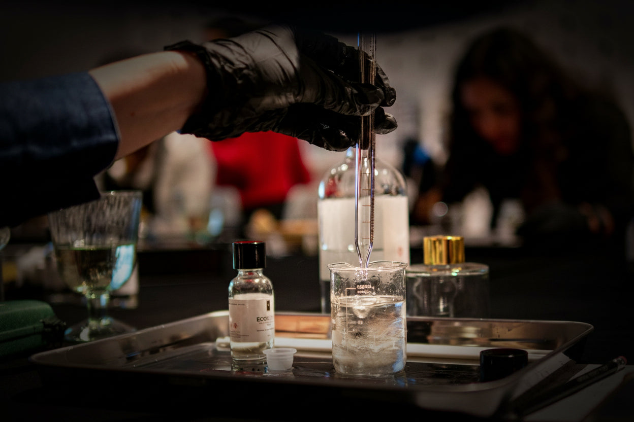 A close-up of a hand wearing a black glove holding a glass dropper over a small bottle on a metal tray at what appears to be a perfume or cosmetic event. A blurred person is visible in the background, and other bottles and glasses are present on a dark table.