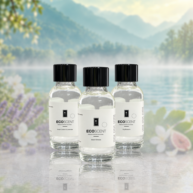 Three small clear glass bottles of EcoScent essential oil have black caps and white labels. They sit on a reflective surface. Behind them is a blurred scenic view of mountains, water, and flowers.