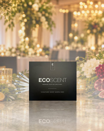 A black ECOSCENT premium eventing scents box is displayed in the foreground, with several white sample strips fanned out to the left. The background is a softly blurred, warm-toned event venue with hanging lights and floral decorations.