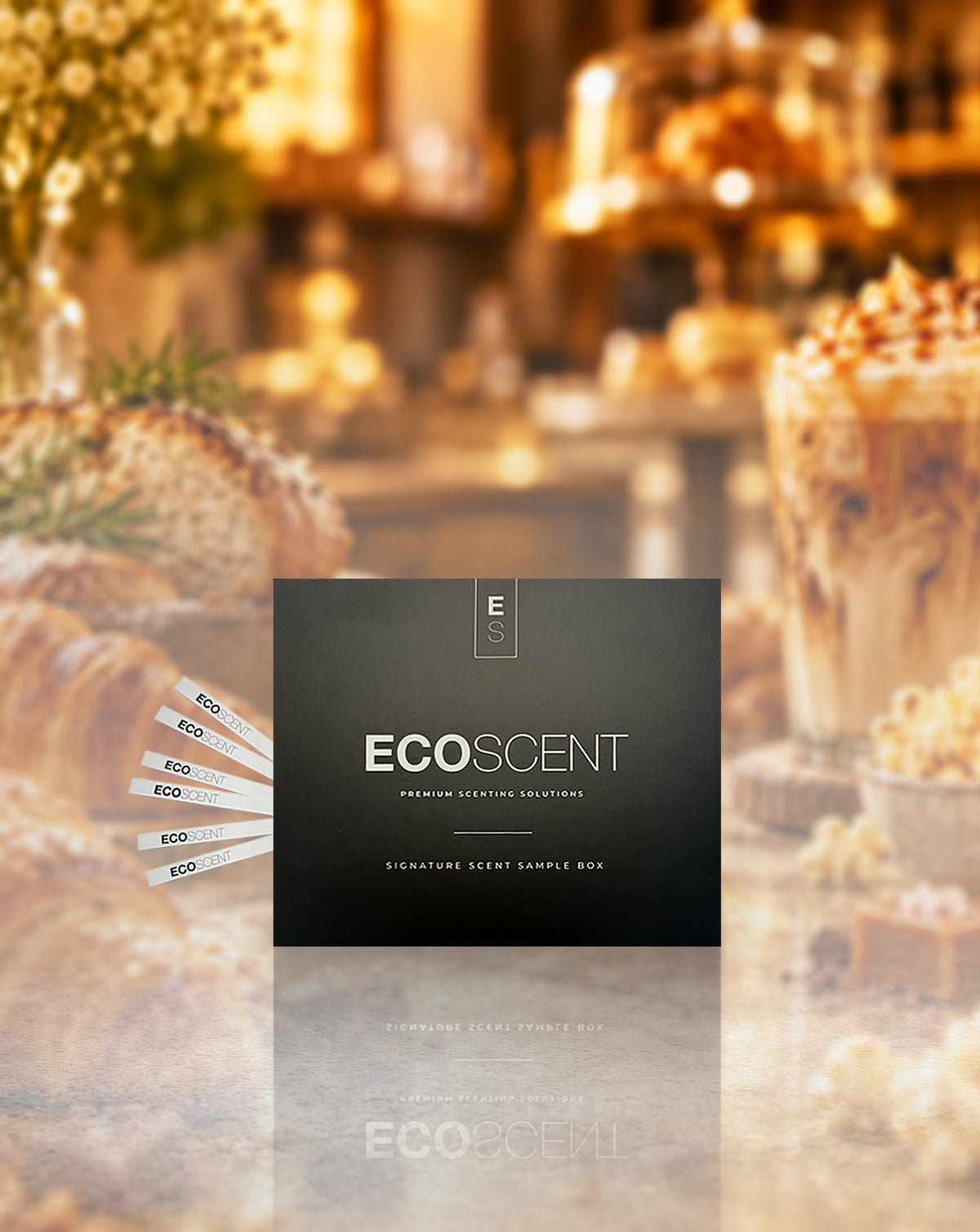 A dark charcoal square product box labeled ECOSCENT is centered over a warm, blurred cafe or bakery scene with pastries and drinks. Several white sample sticks are fanned out to the left.