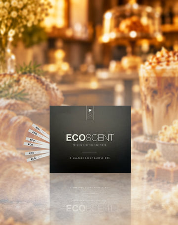 A dark charcoal square product box labeled ECOSCENT is centered over a warm, blurred cafe or bakery scene with pastries and drinks. Several white sample sticks are fanned out to the left.