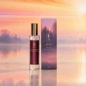 A bottle of Golden Serenity room spray and its matching outer box, both featuring a gradient design with a forest silhouette, set against a warm sunset landscape over a calm lake.