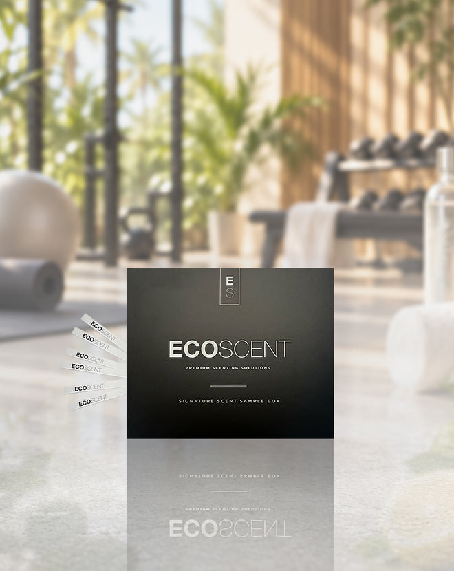 A black ECOSCENT premium setting solution sample box stands on a reflective surface in a softly blurred gym environment with dumbbells, towels, and greenery in the background. Several white sample strips are fanned out to the left.