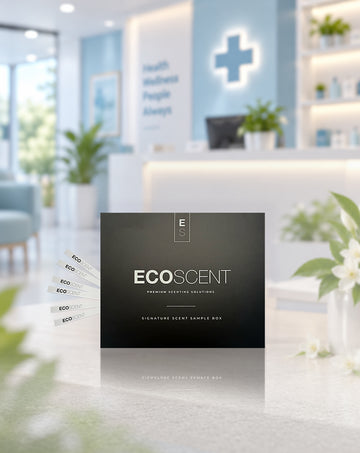 A black square sample box labelled ECOSCENT is centred on a light countertop, with several white sample strips fanned out to the left. The background shows a softly blurred modern clinic or lobby interior with blue walls, a white reception desk, and green plants.