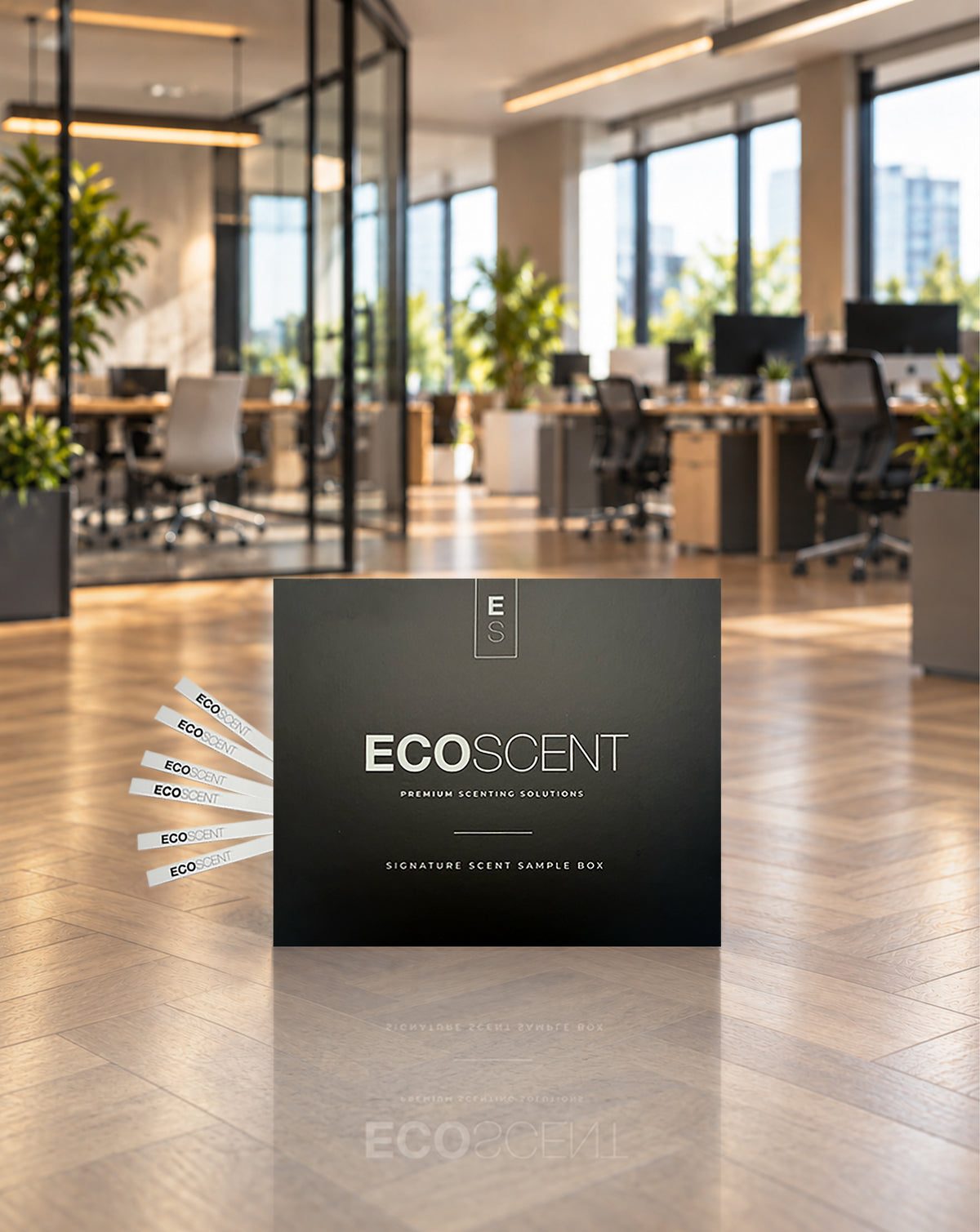 A black ECOSCENT premium office scent sample box stands on a polished wood floor in a blurred modern office space. Several white sample strips are fanned out to the left, and the brand name ECOSCENT is clearly printed on the front.