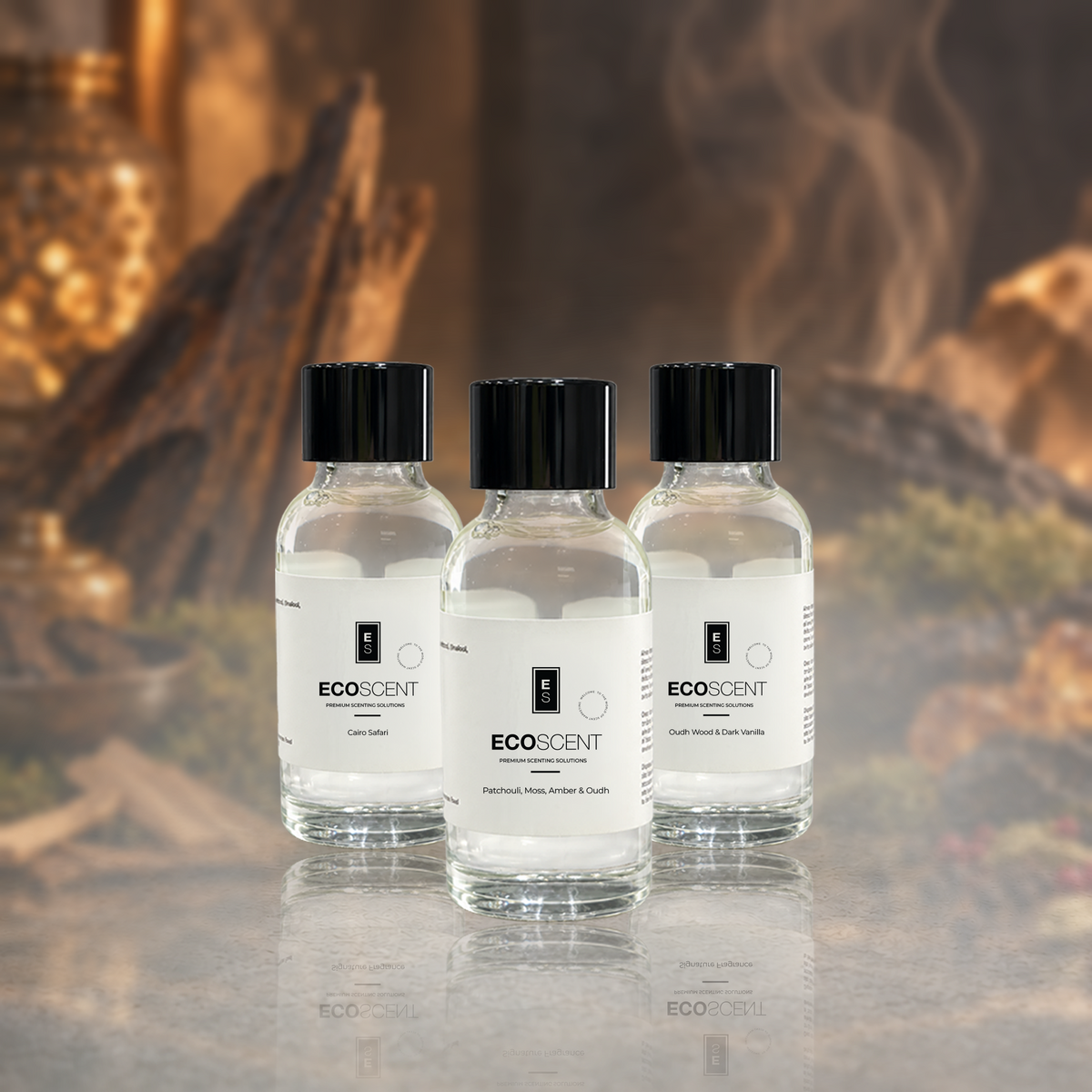 Three small clear glass bottles of EcoScent essential oil with black caps and white labels, arranged on a reflective surface against a warm, smoky background.