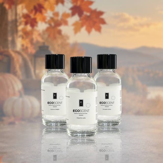 Three clear glass bottles of EcoScent fragrance oil have black caps and white labels.They sit on a reflective surface.A blurred outdoor autumn background is behind them.