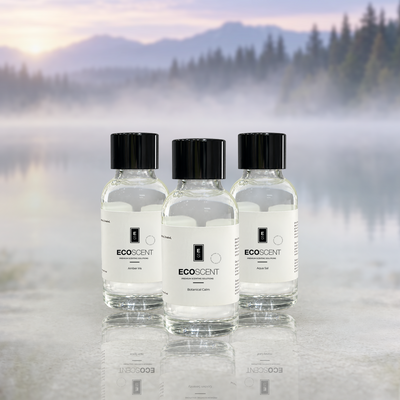 Three clear glass bottles of EcoScent fragrance oils have black caps and white labels. They are arranged on a reflective surface. A misty mountain lake forms the background.