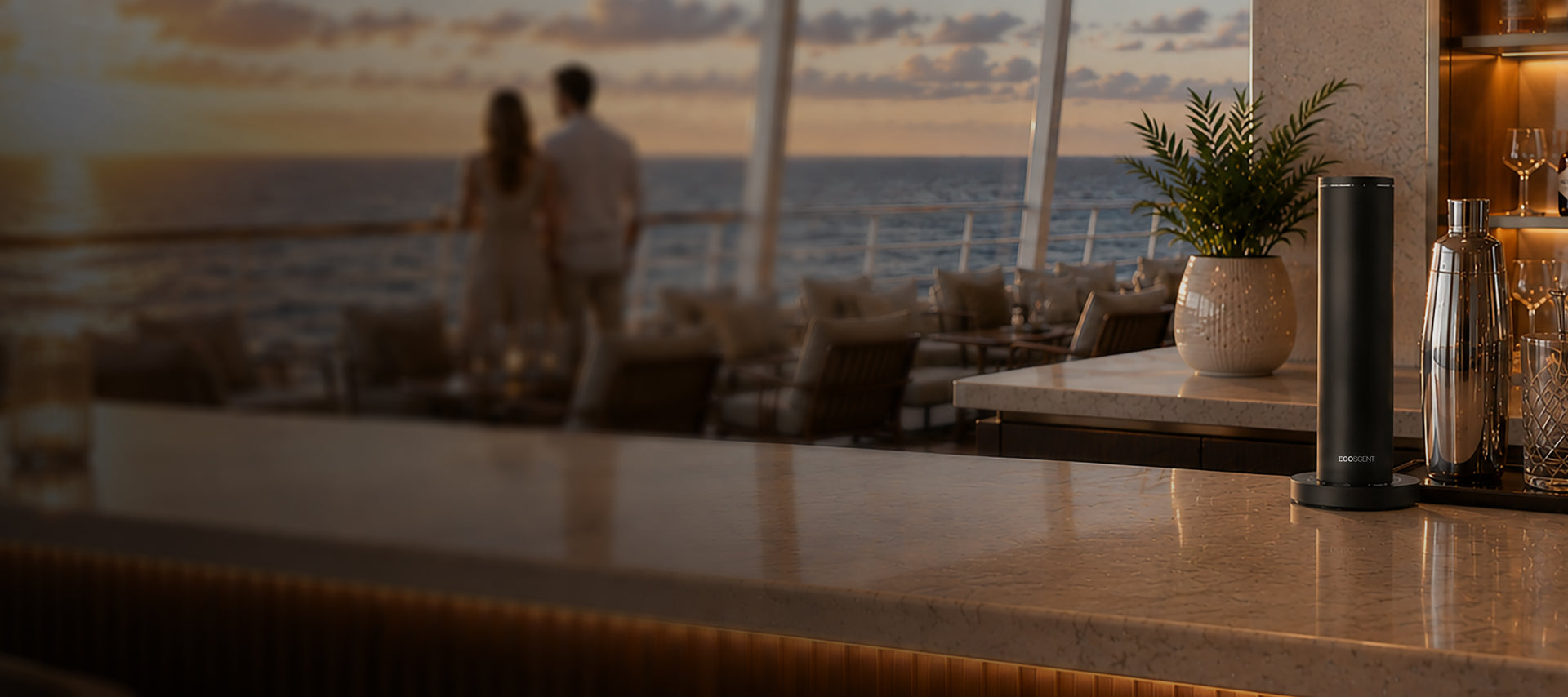 Luxury cruise bar with marble countertop, cocktail shaker, and glassware, overlooking ocean view at sunset with couple in the background, evoking relaxation and elegance.