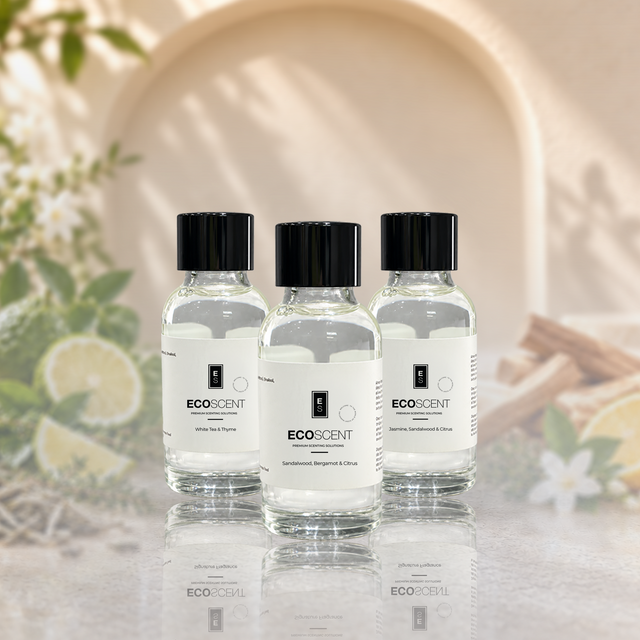 Three small clear glass bottles of EcoScent fragrance oil have black caps. They sit on a reflective surface. The background is warm beige. Soft floral and botanical props surround them.
