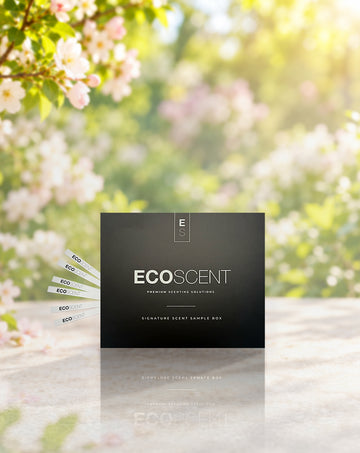 A black EcoScent premium scenting solution box sits on a light stone surface with cherry blossom flowers and green leaves blurred in the background. Several white sample strips are fanned out to the left of the box.