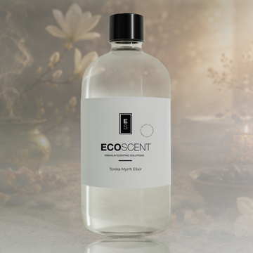 A clear glass bottle of EcoScent Tonka Myrrh Elixir with a white label and black cap, set against a warm, softly blurred background with decorative flowers and candles.