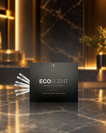 A dark charcoal grey square product box labelled EcoScent sits on a polished marble surface with a blurred luxury interior in the background. Several white sample strips are fanned out to the left of the box.