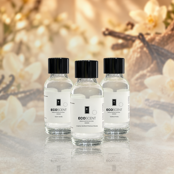 Three small clear glass bottles of EcoScent Vanilla fragrance oil sit in a row. They rest on a reflective surface. A warm, blurred background shows flowers and vanilla beans.