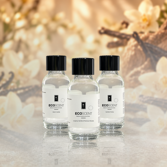 Three small clear glass bottles of EcoScent Vanilla fragrance oil sit in a row. They rest on a reflective surface. A warm, blurred background shows flowers and vanilla beans.