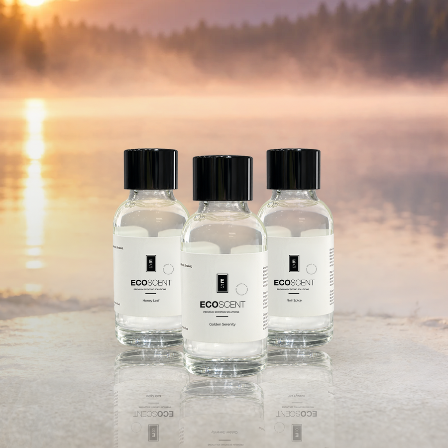 Three clear glass bottles of EcoScent fragrance oil have black caps and white labels. They sit on a reflective surface. A misty lake at sunset fills the background.