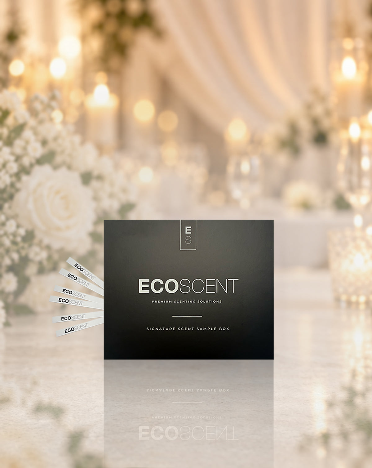 EcoScent Wedding Signature Scent Sample Box.Several white sample sticks fan out to the left.The background is softly blurred and warm.It shows a wedding-style event with lights and flowers.