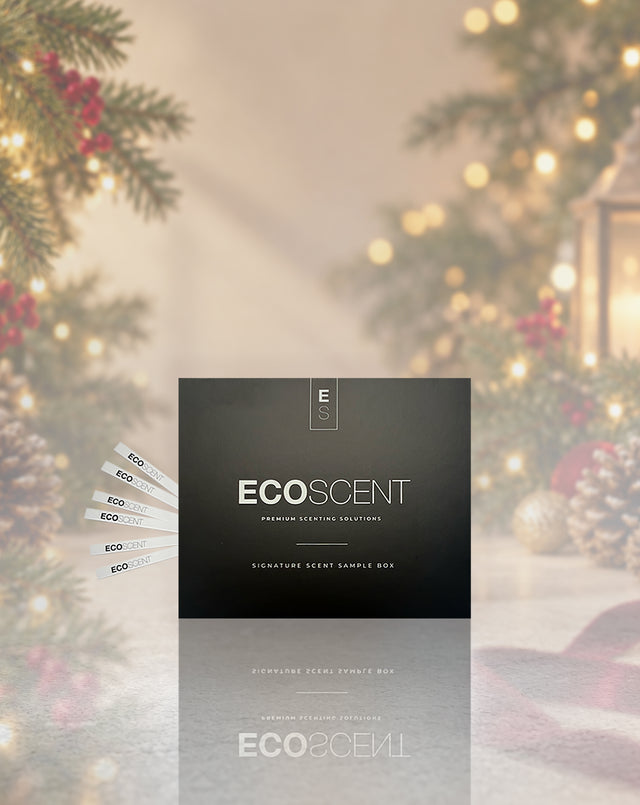A dark charcoal gray ECOSCENT sample box stands upright on a reflective surface, with several white sample strips fanned out to the left. The background is a softly blurred holiday scene with pine branches, red berries, and warm string lights.