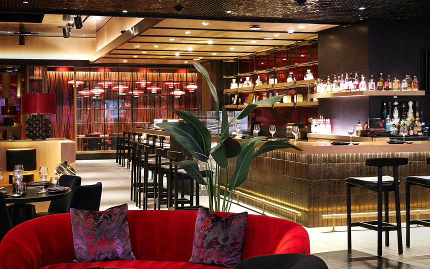 Modern restaurant interior with a stylish bar, tall black chairs, tables set for dining, red velvet couches, decorative pillows, and a large green plant in the foreground. Warm lighting and bottles line the bar shelves.