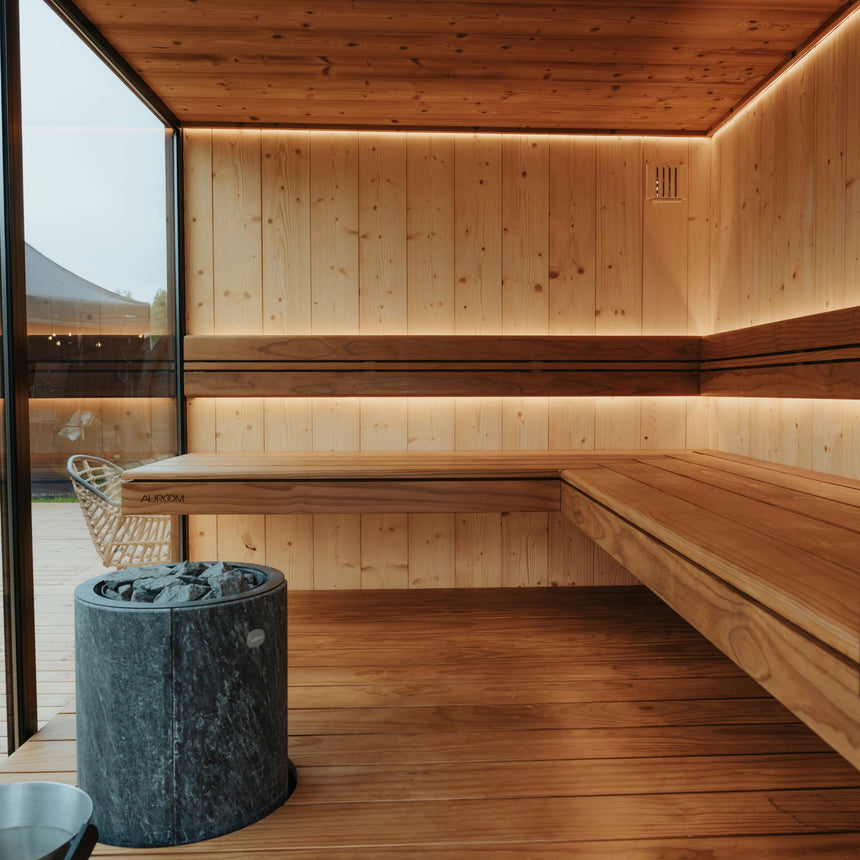 A modern wooden sauna interior with built-in benches, warm ambient lighting, a stone sauna stove, and glass doors opening to an outdoor deck.