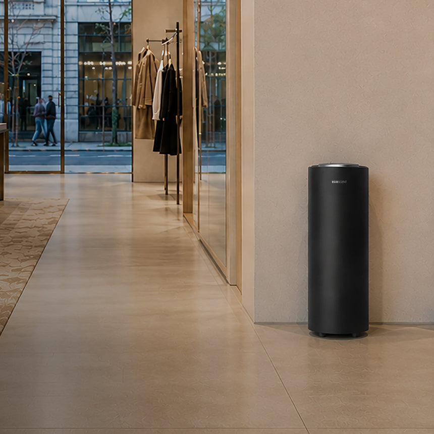 A sleek, matte black cylindrical standalone scent machine is positioned against a beige wall in a modern, minimalist boutique interior with warm lighting and large windows.