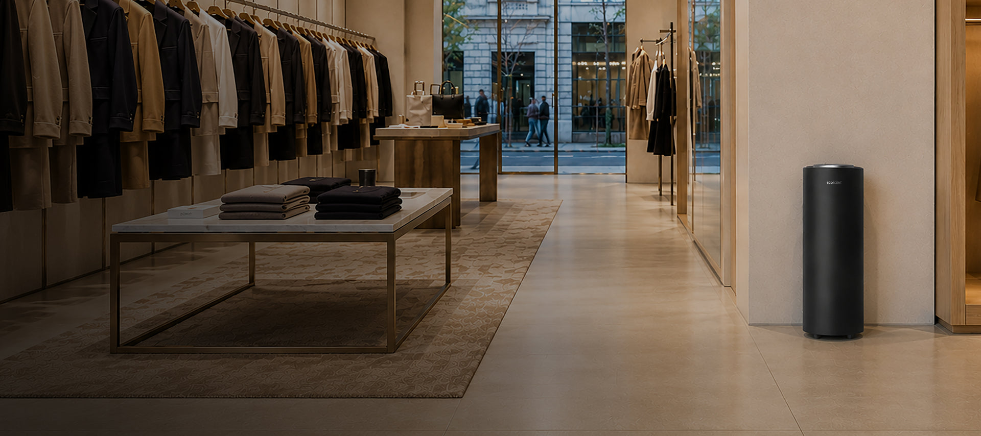 A sleek, matte black cylindrical floor scent machine stands on a light beige tiled floor in a modern, minimalist clothing boutique.