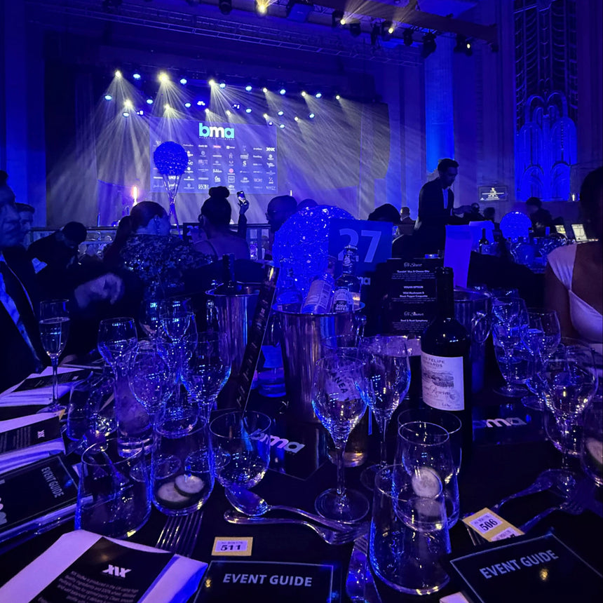 A dimly lit event hall with blue stage lighting, tables set with wine glasses and menus, and a large 'bma' stage backdrop in the distance.